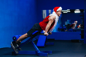 Holidays and celebrations, New year, Christmas, sports, bodybuilding, healthy lifestyle - Muscular handsome sexy Santa Claus. Young handsome fit man performing hyperextension exercise on roman chair