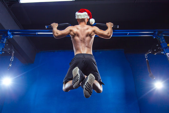 Holidays and celebrations, New year, Christmas, sports, bodybuilding, healthy lifestyle - Muscular handsome sexy Santa Claus. Athlete muscular fitness male model pulling up on horizontal bar in a gym