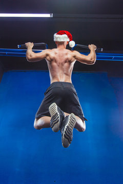 Holidays and celebrations, New year, Christmas, sports, bodybuilding, healthy lifestyle - Muscular handsome sexy Santa Claus. Athlete muscular fitness male model pulling up on horizontal bar in a gym
