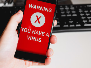 Hand holding a phone with virus alert on screen with keyboard. Virus concept. First person point of...