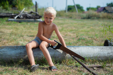 young boy without a t-shirt, sitting on a tube sawing a branch with a saw in the countryside.