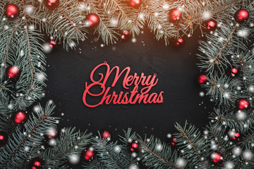 Black stone background with a frame of fir branches decorated with balls and red ribbon. Top view. With Merry Christmas text. Snow effect