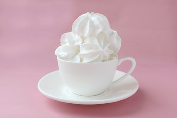Side view on white meringues in a white teacup on a white plate on a pink background. Copy space