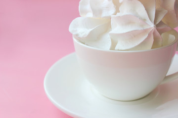 Side view on white meringues in a white teacup on a white plate on a pink background. Copy space