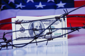 US Dollar money wrapped in barbed wire against United States national flag as symbol of economic...