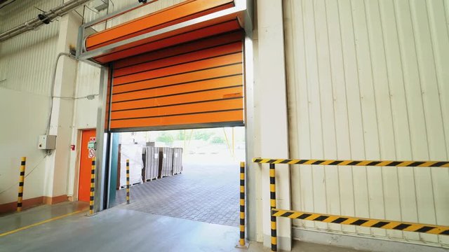 automatic roller gates of orange color are lowering down in the warehouse of woodworking plant. Camera movement to the right