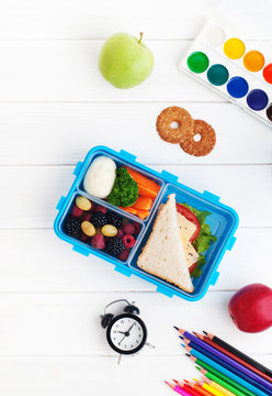 Lunch box with sandwich, vegetables, berries on white wooden background with clock and school accessories