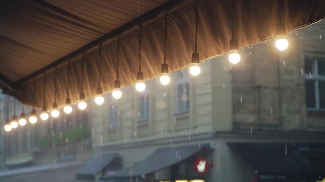 decorative lamps are hanging in the row and lighting on the background of the rain. Raindrops are falling on the roof of the summer terrace of a cafe which is decorated with a row of lanterns