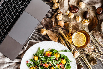 Desktop flat lay. Laptop, Salad with mango, arugula, pomegranate seeds lying on white plate. Next lying pumpkin, cup of black tea with lemon, dry flowers and leaves, physalis, chestnuts, sweater