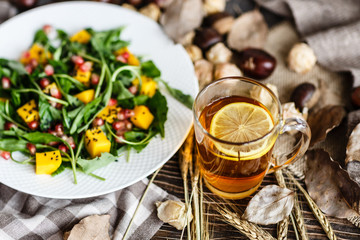 Food flat lay. Top view. Salad with mango, arugula, pomegranate seeds lying on white plate. Next lying pumpkin, cup of black tea with lemon, dry flowers and leaves, physalis, chestnuts, sweater