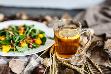 Salad with mango, arugula, pomegranate seeds lying on white plate. Next lying pumpkin, cup of black tea with lemon, dry flowers and leaves, physalis, chestnuts, sweater