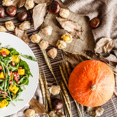 Food flat lay. Top view. Salad with mango, roasted pumpkin, arugula, pomegranate seeds lying on white plate. Next lying dry flowers and leaves, physalis, chestnuts