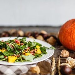Salad with mango, roasted pumpkin, arugula, pomegranate seeds lying on white plate. Next lying dry flowers and leaves, physalis, chestnuts
