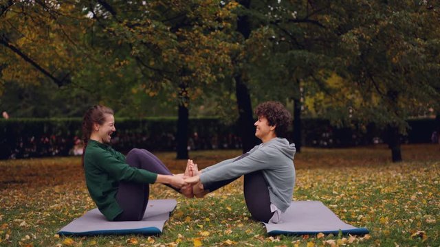 Happy young women are doing pair yoga having fun and laughing sitting on mats on grass in park. Beautiful autumn nature lawn and trees are in background.