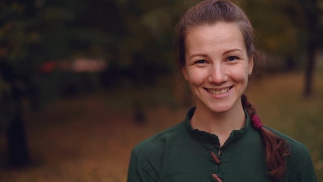 Close-up portrait of beautiful girl with braid looking at camera and smiling then laughing standing outdoors in park in autumn with trees in background.