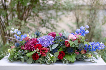 Elongated floral arrangement in vintage metal vase. table setting. Blue and red color. Gorgeous bouquet of different flowers. Bright room, stucco on the wall