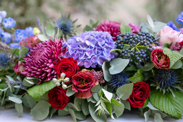 Elongated floral arrangement in vintage metal vase. table setting. Blue and red color. Gorgeous bouquet of different flowers. Bright room, stucco on the wall