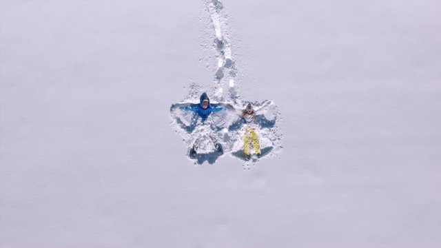 Aerial - Couple hold hands, fall on their backs and start making snow angels