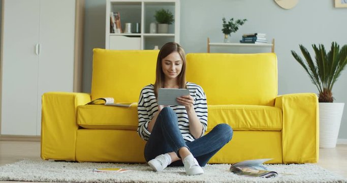 Young beutiful smiled woman sitting at home on the floor in the living room with a tablet device and taping on its screen while watching something.