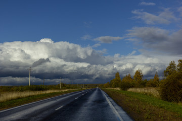 asphalt road among the fields and forests illuminated by the sun, the sky with textured clouds and clouds and a rainbow