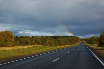 asphalt road among the fields and forests illuminated by the sun, the sky with textured clouds and clouds and a rainbow