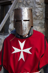 Obraz premium stylized costume clothes of a knight in a helmet in a red cloak with a cross and a sword on the background of an ancient stone fortress