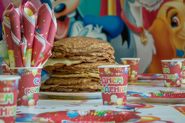 A large portion of pancakes on the festive table of the children's birthday. Disposable holiday dishes in Burgundy color and napkins folded in a tube.