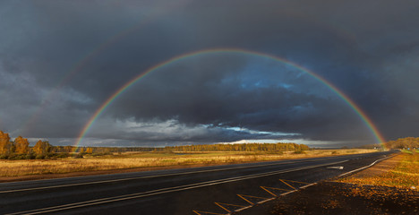 panoramic shot of a full double rainbow, against a dark storm sky, surrounded by Golden fields and the road going away