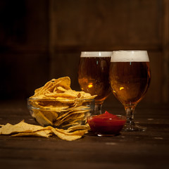 Two glasses with a light beer. Next to the bowl with nachos and sauce bowl with tomato sauce. On a dark wooden table.