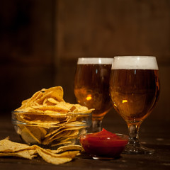 Two glasses with a light beer. Next to the bowl with nachos and sauce bowl with tomato sauce. On a dark wooden table.