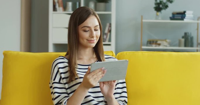 Portrait of the charming young woman in the striped blouse chatting and scrolling on the tablet device with a smile.