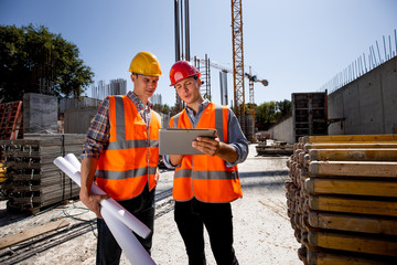 Architect  and structural engineer dressed in orange work vests and  helmets discuss a building...