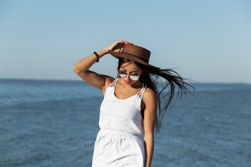 Fototapeta na wymiar Portrait of stunning dark-haired girl in white dress, sunglasses and brown hat near the sea on a sunny day.