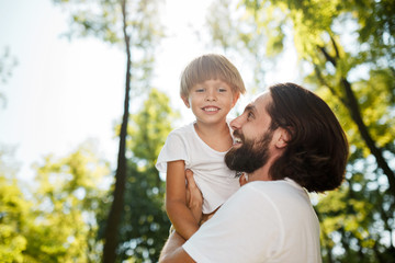 Handsome dark-haired father with beard dressed in the white t-shirt is holding in the arms his little son, watching at him and smiling in the park.