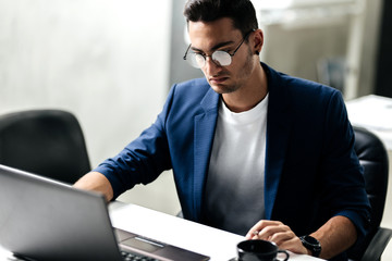 Dark-haired young architect in glasses dressed in blue checkered jacket  works on the laptop in the office