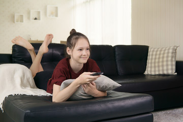Obraz premium cute preteen girl watching TV on couch using remote control. Living room interior in background. Text space