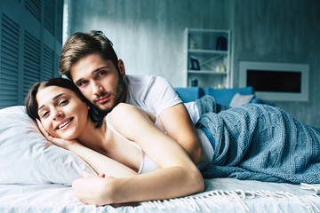 Happy beautiful young couple are lying, hugging and kissing in a comfortable bed at home. Love emotions