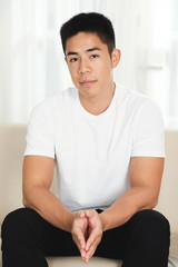 Attractive unsmiling mixed-race man sitting on couch and looking at camera
