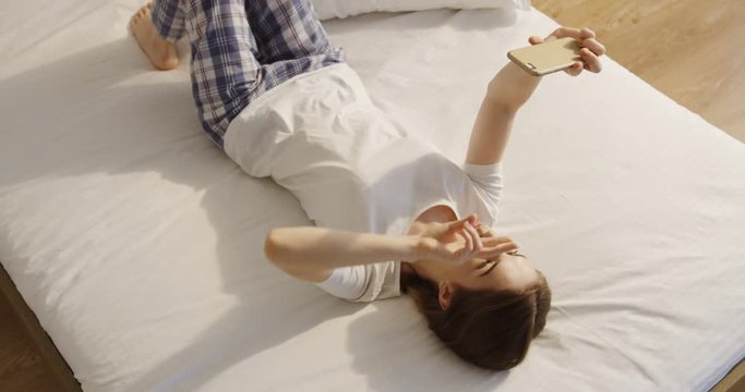 View from above on the attractive Caucasian young woman in pajama lying on the bed and taking selphie pictures on the smartphone camera.