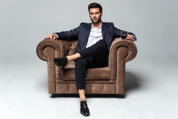 Little stylish rest. Full length of handsome young man looking at camera while sitting against white background in big chair - Powered by Adobe