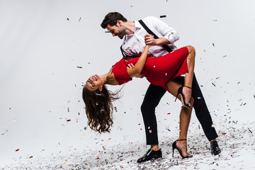 It is hot salsa! Full length of young beautiful couple dancing while standing against white background with confetti  - Powered by Adobe