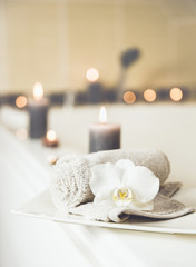 Small rolled towel with white orchid blossom on white ceramic tray, burning candles on the background in bath room. Fresh dry clean laundry concept.