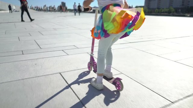A little girl in an iridescent bright skirt is riding a scooter in the city. Slow motion