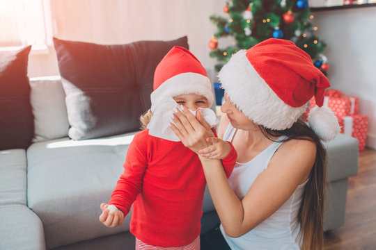 Funny photo of woman sits besides her child and covers her mouth with white napkin. Family is sick at Christmas. They have handkerchief. Sick people have runny nose. T She wears red Christmas hat. And