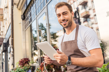 smiling handsome florist holding tablet near flower shop and looking at camera