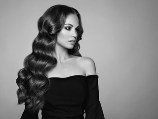 Fototapeta premium Brunette Girl with Long Healthy and Shiny Curly Hair. Care and Beauty. Beautiful Model Woman with Wavy Hairstyle. Make-Up and Black Dress