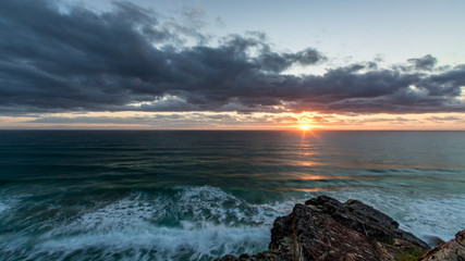 Sun rising through cloudy sky over ocean and rocky coastline
