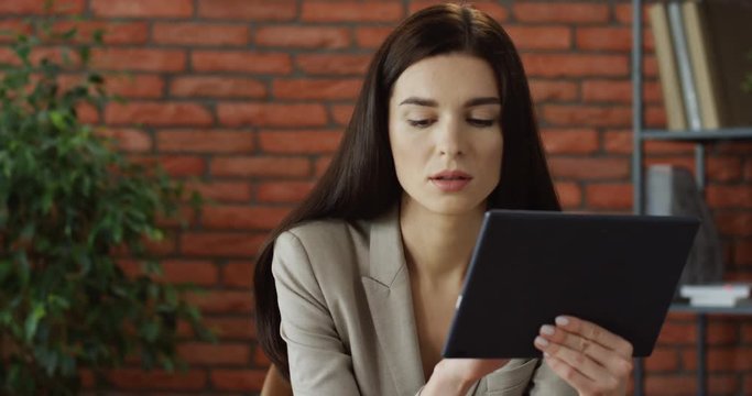 Young pretty brunette woman sitting in her office and using tablet computer, then looking at the screen and smiling. Close up.
