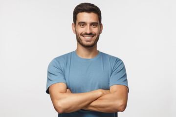 Portrait of smiling handsome man in blue t-shirt standing with crossed arms isolated on grey...
