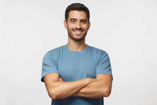 Indoor portrait of young european caucasian man isolated on gray background, standing in blue t-shirt with  crossed arms, smiling and  looking straight at camera